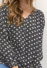 Kobieta ubrana w czarną i białą wzorzystą bluzkę z dekoltem w serek i długimi rękawami, siedząca na jasnym tle.