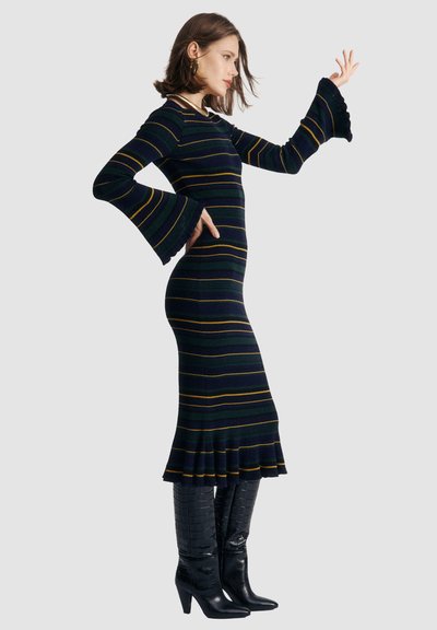 Juostuotas megztinis suknelė tamsiai žalia ir tamsiai mėlyna, su išplatintomis rankovėmis ir klostuotu apvadu. Dėvima su juodomis batu kompiuterių aukštakulniams su krokodilo raštu.