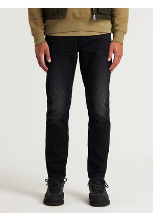 Homme portant un jean noir et des bottes noires à lacets, debout devant un fond clair uni.