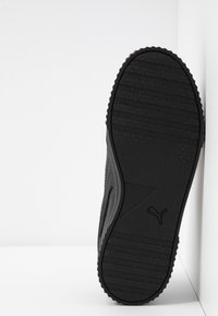 Semelle de chaussure de sport noire en caoutchouc texturé, dotée de rainures et d'une zone lisse près de la voûte plantaire, marquée d'un logo subtil.
