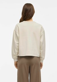 Svetlo bež skrajšan sweatshirt z dolgimi rokavi, rebrastimi manšetami in okroglo ovratnikom, nosi pa se ga over rjavimi širokimi hlačami. Gladka tekstura.