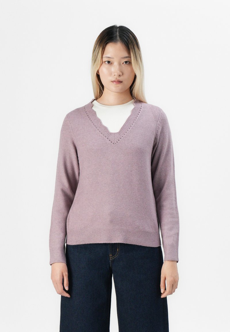 Jovem mulher com cabelo loiro liso usa um suéter V-de decote leve em roxo sobre uma blusa branca e calças largas de tom azul escuro, em pé contra um fundo branco.