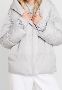 Světlá šedá péřová bunda s velkým límcem, prošívanou strukturou a předními kapsami, vrstvená přes šedý svetr.