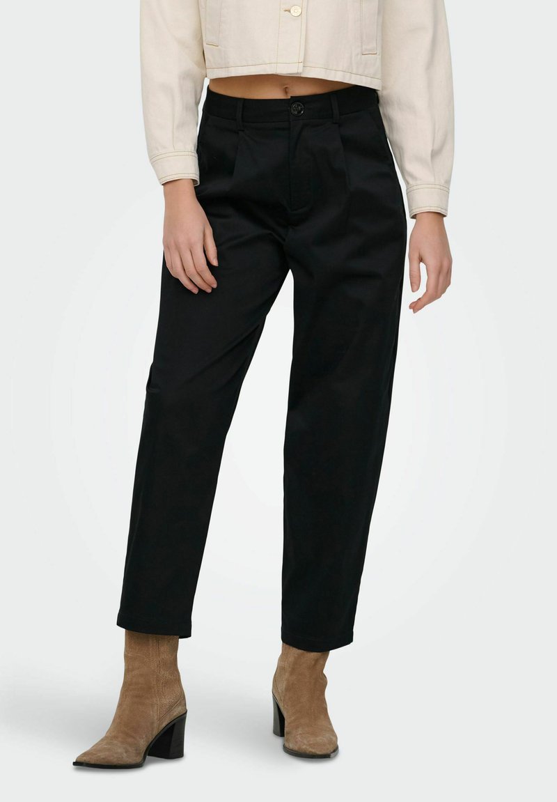 Femme portant un pantalon noir taille haute associé à des bottines à talons en daim beige et une veste courte beige, sur un fond uni.