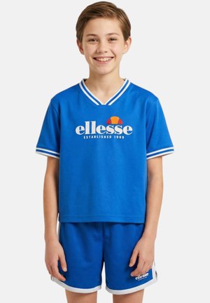 Garçon souriant portant un t-shirt de sport ellesse bleu et un short assorti avec bordure blanche, debout devant un fond uni.