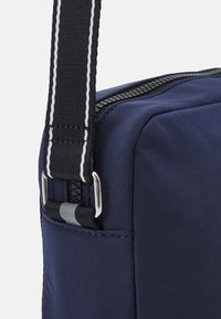 Angolo di una borsa in tessuto blu navy con manico regolabile a strisce bianche e nere e zipper nera visibile.