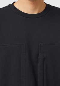 Felpa nera con scollo rotondo, caratterizzata da due tasche rettangolari frontali, realizzata in un tessuto liscio di peso medio.