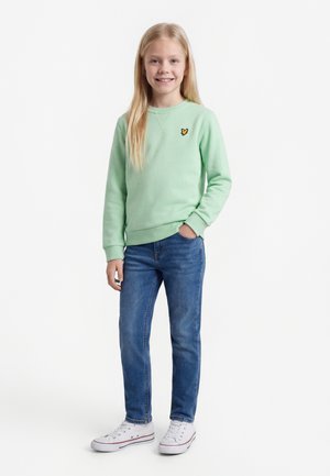 Młoda dziewczyna z długimi blond włosami, ubrana w jasną zieloną bluzę, niebieskie dżinsy i białe tenisówki, uśmiechająca się z jedną ręką w kieszeni.