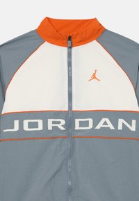 Giacca leggera con zip nei colori grigio, arancione e bianco. Presenta un colletto rialzato, il testo 'JORDAN' in bold e un logo arancione sul petto.