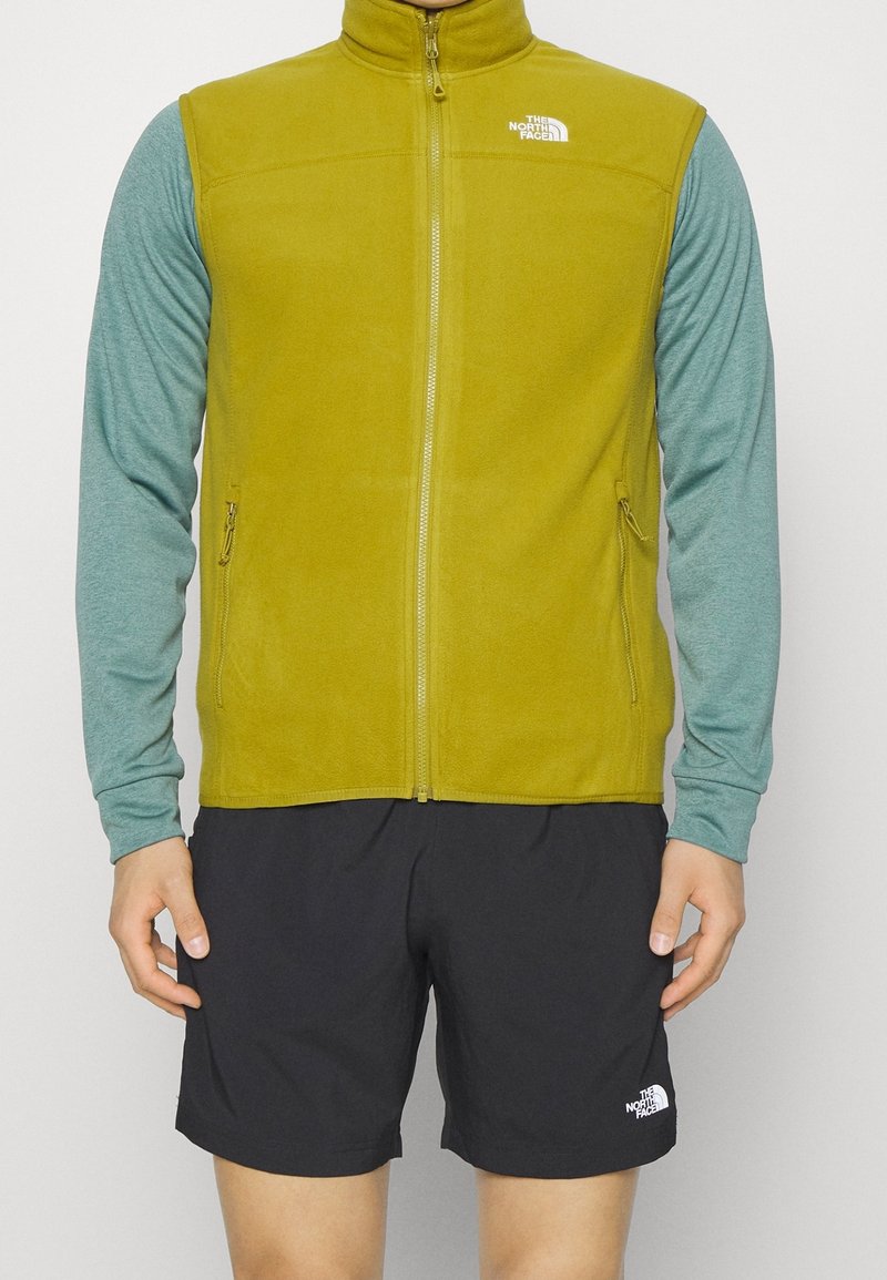 Olivgrüne Fleece-Weste mit hohem Kragen, Reißverschlusstaschen und einem kleinen Logo. Getragen über einem hellen teal-farbenen Langarmshirt und schwarzen Shorts.