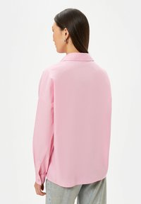 Camicia rosa a maniche lunghe con colletto, texture liscia e polsini abbottonati. Taglio morbido e orlo arrotondato. Indossata con jeans di denim chiaro.