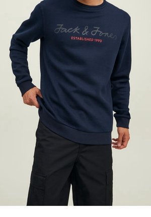 Laivastonsininen collegepaita, jossa on "Jack & Jones Established 1990" -teksti, yhdistetty mustiin cargo-housuihin, kädet säätämässä collegepaidan helmaa.