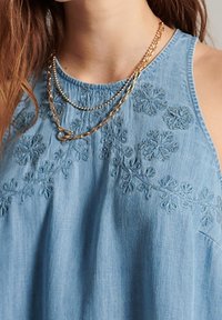 Top sin mangas de denim con bordado floral en tonos azules, de corte relajado y fabricado en una textura suave. Collares en capas doradas complementan el conjunto.
