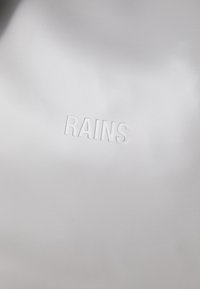 Una superficie grigia chiara e liscia presenta in modo prominente la parola "RAINS" in rilievo al centro in basso. Lo sfondo è minimalista.