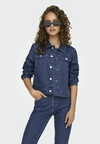 Giacca in denim blu scuro con tasche frontali, chiusura a bottoni e maniche con risvolto. Indossata con jeans in denim abbinati. Texture liscia, design casual.