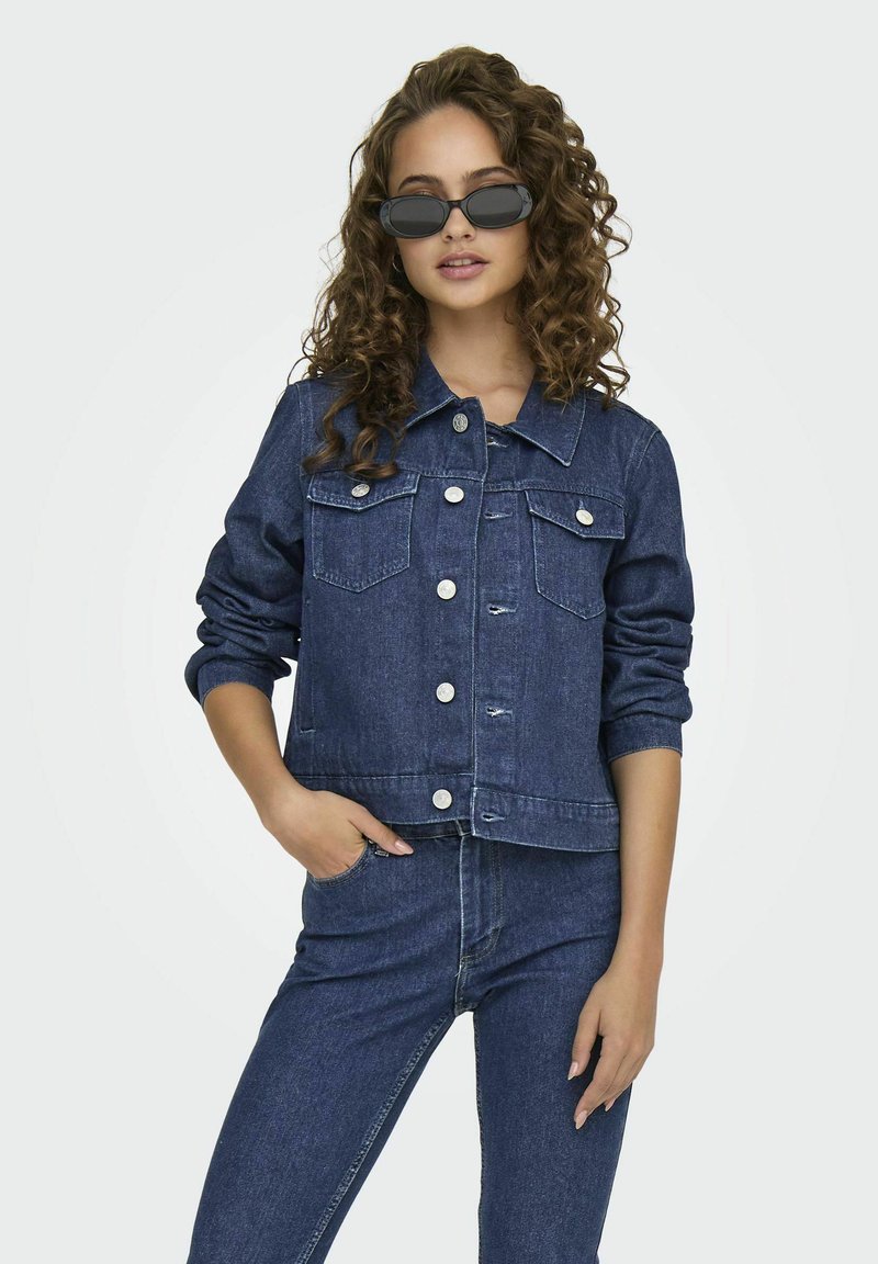 Giacca in denim blu scuro con tasche frontali, chiusura a bottoni e maniche con risvolto. Indossata con jeans in denim abbinati. Texture liscia, design casual.