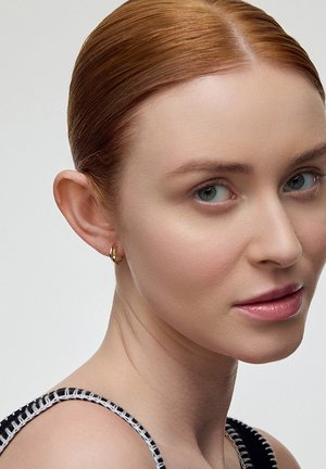 Złota kolczyk w kształcie koła na modelce o jasnej karnacji i rudych włosach. Tło jest jednolite, skupiając się na kolczyku i cechach twarzy.