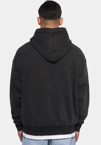 Czarna bluza z kapturem, z regulowanym sznurkiem, w luźnym kroju, z prążkowanymi mankietami i dołem. Miękka tekstura, minimalistyczny design, noszona z jasnoniebieskimi dżinsami.