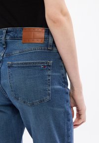 Kék farmernadrág magas derékkal, barna bőr címke a "TOMMY JEANS" felirattal és egy kis logófelvarróval a hátsó zseben.