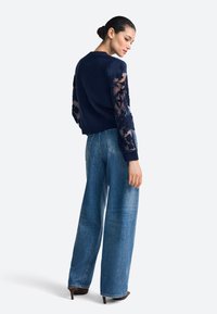 Maglione blu navy con maniche in mesh floreale texturizzato, abbinato a jeans a gamba larga blu e tacchi. Il maglione presenta una scollatura arrotondata e un orlo a coste.