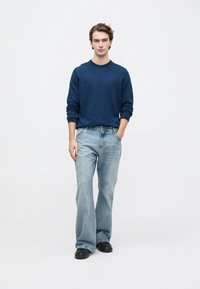 Νεαρός άνδρας που στέκεται με το ένα χέρι στην τσέπη, φορώντας μπλέ πουλόβερ, ανοιχτό μπλε καμπάνα τζιν και μαύρα παπούτσια σε λευκό φόντο.