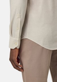 Camisa de manga larga color beige claro con una textura suave, puños de botón único y dobladillo curvado, combinada con pantalones de tela en color topo de corte ajustado.
