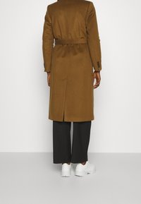 Cappotto marrone in misto lana con cintura, spacco posteriore e polsini con bottoni, abbinato a pantaloni scuri e sneakers bianche. Presenta una texture liscia.