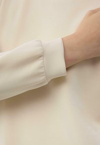 Maglia a maniche lunghe di colore beige chiaro, con polsino arricciato e texture del tessuto liscia. Design semplice senza motivi o accenti.