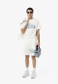 Conjunto de camiseta de algodón blanca y pantalones cortos con un logo gráfico azul. Sosteniendo una bolsa gris, lleva gafas de sol y zapatillas blancas. Corte casual.