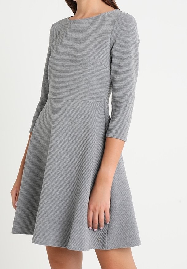 Femme portant une robe en maille côtelée grise, longueur genou, avec des manches trois-quarts et une taille cintrée, debout devant un fond uni.