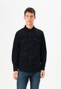 Camisa negra de botones en tejido texturizado con dos bolsillos en el pecho y costuras en contraste. Combinada con vaqueros azules, mostrando un corte recto.