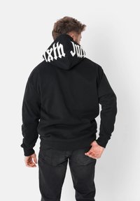 Svart hettegenser laget av mykt stoff, med stor grafisk tekst i hvitt på hetta. Kenguru-lomme og ribbestrikkede mansjetter og nederkant.