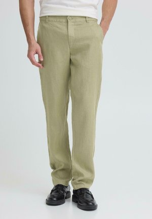 Mann trägt hellolivgrüne Hose mit geradem Bein, weißes Hemd und schwarze Leder-Loafer, steht mit den Händen in den Taschen.