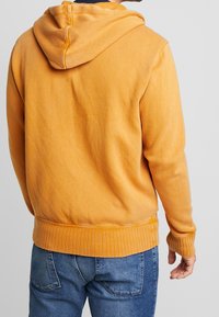 Sudadera recortada en color mostaza suave, confeccionada con tela blanda. Presenta un cinturón y puños acanalados, con capucha ajustable.