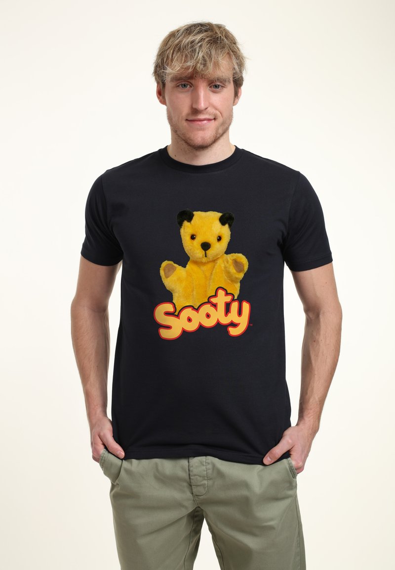 Svart t-shirt med kort ärm som visar en grafisk av en gul plyschbjörn med svarta öron, som håller upp sina armar, och ordet "Sooty" i orange nedanför.