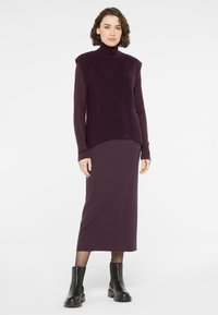 Lila ärmelloses Rollkragenoberteil aus strukturiertem Strick, kombiniert mit einem figurbetonten lila Rock. Schwarze Knöchelstiefel runden das Outfit ab.