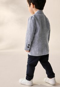 Hellblaues Leinenblazer mit dunklen Knöpfen, kombiniert mit marineblauen Hosen und weißen Sneakers. Das Outfit zeichnet sich durch ein strukturiertes Design und umgeschlagene Ärmel aus.