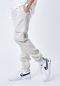 Pantaloni cargo beige chiaro con polsini elasticizzati, tasche laterali e texture liscia. Abbinati a sneakers bianche con dettagli neri.