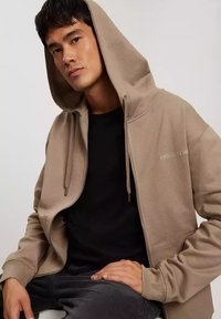 Beżowa bluza z kapturem i zamkiem, z sznurkiem, ściągaczem na rękawach i przednimi kieszeniami. Czarny T-shirt pod spodem.