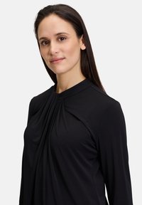 Svart långärmad topp i slät tyg, med hög halsringning och detaljer med veck fram, designad för en strömlinjeformad passform.