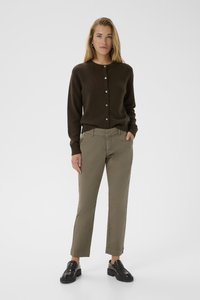 Cardigan a maglia marrone con chiusura a bottoni, abbinato a pantaloni verde oliva e mocassini neri. Il modello sta in piedi con una mano in tasca.