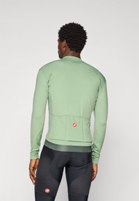 Long-sleeve cycling jersey in light green fabric with a fitted design, featuring a rear pocket and branding detail at the back hem.