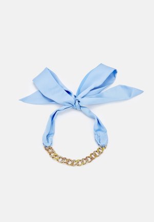 Vaaleansininen kangashuivi, joka on solmittu rusetiksi ja kiinnitetty lyhyeen kultaketjuun, jossa on suuret lenkit, valkoisella taustalla.