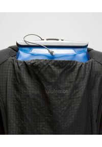 Mochila negra con una textura a cuadros, que sostiene un recipiente de agua azul con un asa gris, mostrando la marca "lululemon" en la tela.