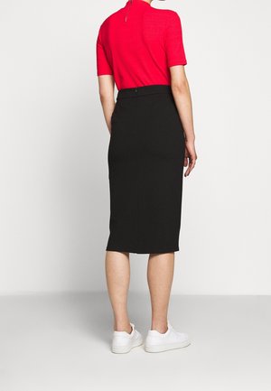 Henkilö, joka pukeutunut punaiseen lyhythihaiseen paitaan, mustaan polvenmittaiseen hameeseen ja valkoisiin tennareihin, seisoo yksinkertaisen vaalean taustan edessä.