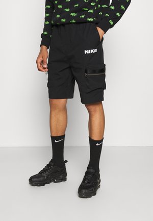 Czarne szorty cargo z elastycznym pasem, kieszenią na suwak i białym logo Nike. Noszone z czarnymi skarpetkami i sportowymi butami Nike.