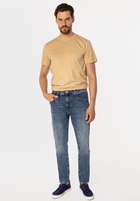 Beżowy bawełniany t-shirt z krótkim rękawem i okrągłym dekoltem; noszony z jasnoniebieskimi dżinsami o prostym kroju, lekko spranym odcieniu oraz granatowymi trampkami z białą podeszwą.