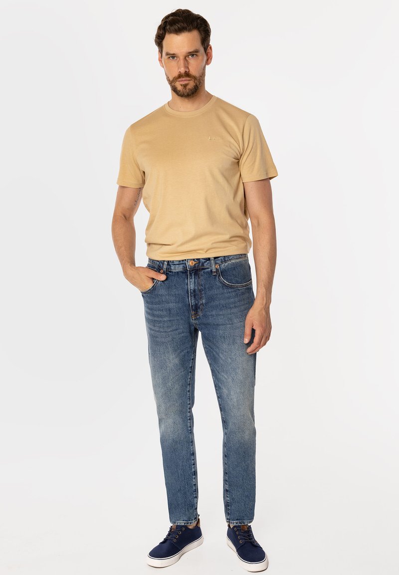 Beżowy bawełniany t-shirt z krótkim rękawem i okrągłym dekoltem; noszony z jasnoniebieskimi dżinsami o prostym kroju, lekko spranym odcieniu oraz granatowymi trampkami z białą podeszwą.