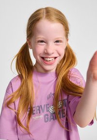 Lila T-Shirt mit einem Volleyball-Graphic und Text, mit kurzen Ärmeln und lockerer Passform. Lange gerade Haare mit Zöpfen.