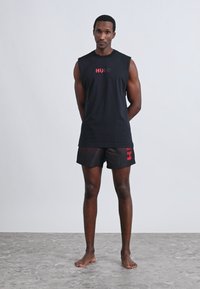 Črna telovadna majica brez rokavov z rdečim napisom "HUGO"; črne kratke hlače z rdečimi poudarki. Model stoji boso na svetlem ozadju.